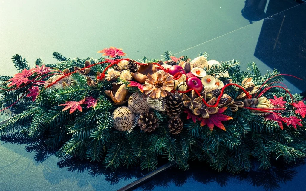 Ozdoby cmentarne powinny być stonowane i stosowne do okazji Ozdobny stroik świąteczny stworzony z gałęzi świerkowych, suchych owoców, szyszek oraz czerwonych i złotych ozdób, starannie ułożony na gładkiej, połyskliwej powierzchni.
