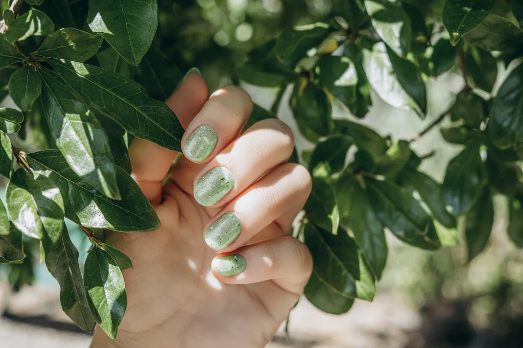 Dłoń z zielonym manicure trzymana wśród gęstych liści drzewa, światło słoneczne prześwieca przez listowie, podkreślając połysk lakieru na paznokciach.