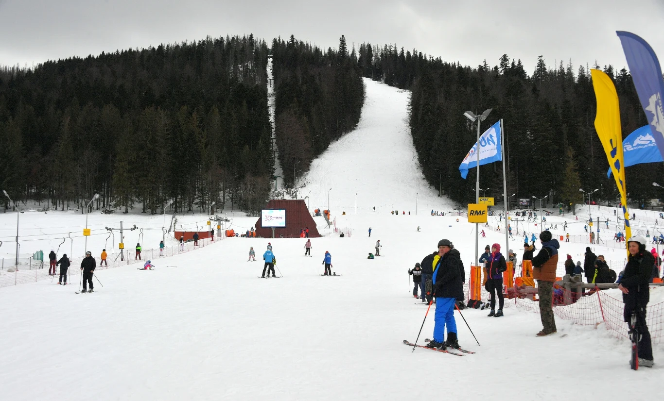 Grupa narciarzy korzystająca z dobrze przygotowanego stoku położonego w otoczeniu lasów, na pierwszym planie osoby z nartami i kijkami, w tle trasa narciarska biegnąca wśród drzew, widoczne również flagi i punkt obsługi przy stoku.