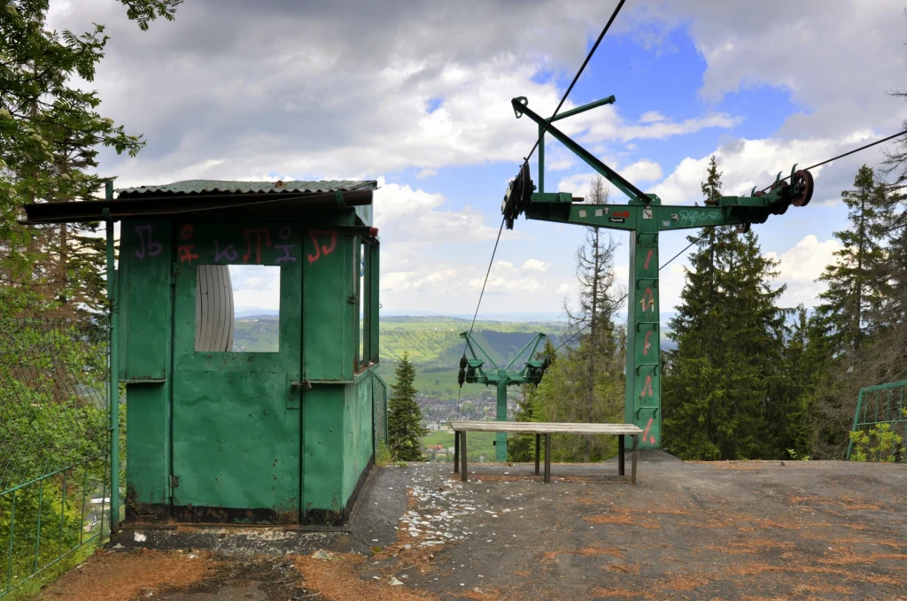Stara zielona budka operatora oraz opuszczona stacja wyciągu krzesełkowego na tle lasu i górskiej panoramy, w otoczeniu porozrzucanych liści i igliwia.