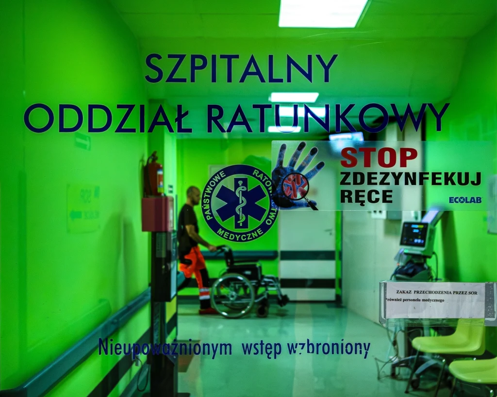 SOR w Szpitalu Mragowskim im. Michala Kajki Korytarz w szpitalnym oddziale ratunkowym o intensywnie zielonych ścianach, widoczny znak zakazu wstępu dla osób nieupoważnionych, plakaty promujące dezynfekcję rąk, osoba siedząca na wózku inwalidzkim oraz pracownik medyczny.