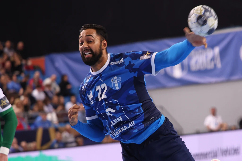 Melvyn Richardson Zawodnik w niebieskim stroju sportowym w trakcie dynamicznego rzutu piłką podczas meczu piłki ręcznej, w tle widoczni kibice oraz fragment boiska.