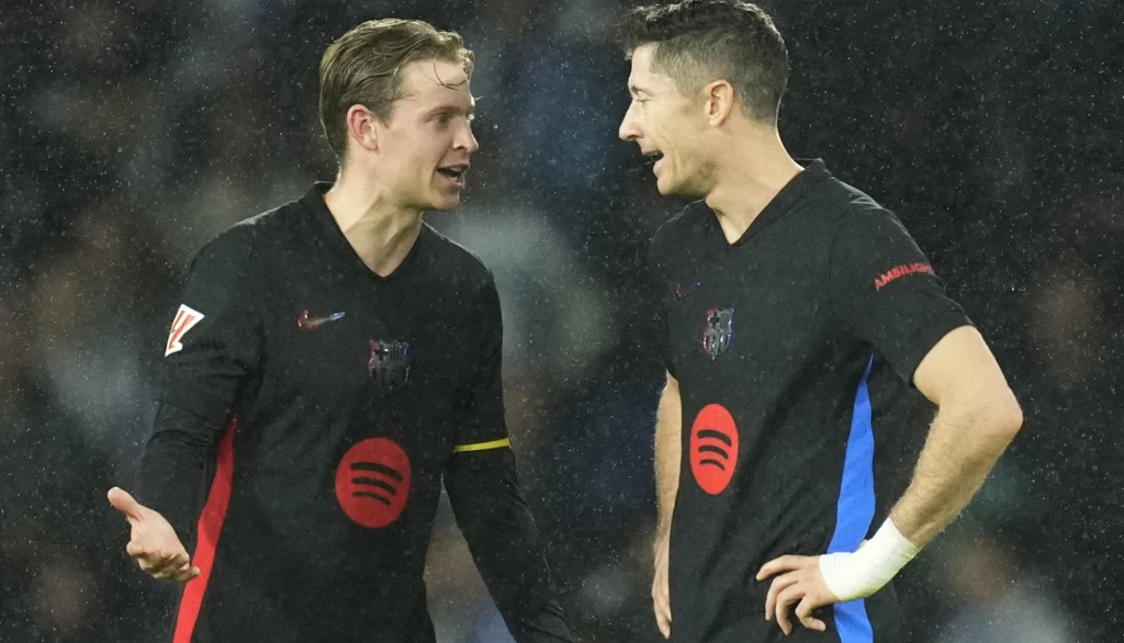 Frenkie de Jong w towarzystwie Roberta Lewandowskiego Dwaj piłkarze drużyny FC Barcelona w czarnych strojach z logo Spotify rozmawiają ze sobą na boisku podczas meczu, sprawiając wrażenie zaangażowanej wymiany zdań, w tle widoczna lekka mgła lub deszcz.