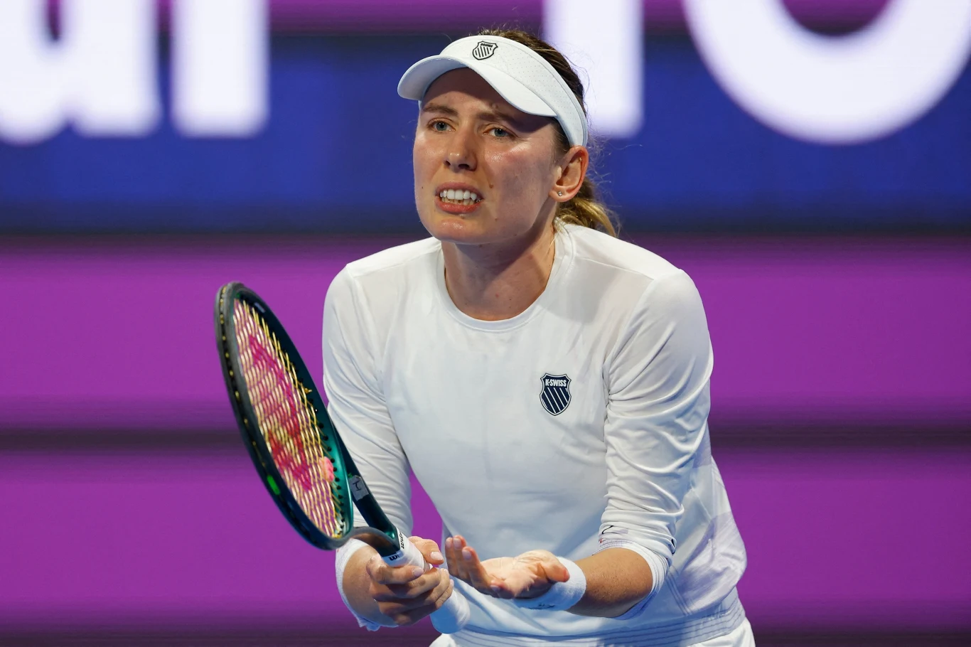 Kobieta w białym stroju sportowym i białej czapce z daszkiem trzyma rakietę tenisową na tle fioletowego kortu, wyrażając emocje mimiką twarzy i gestami rąk.