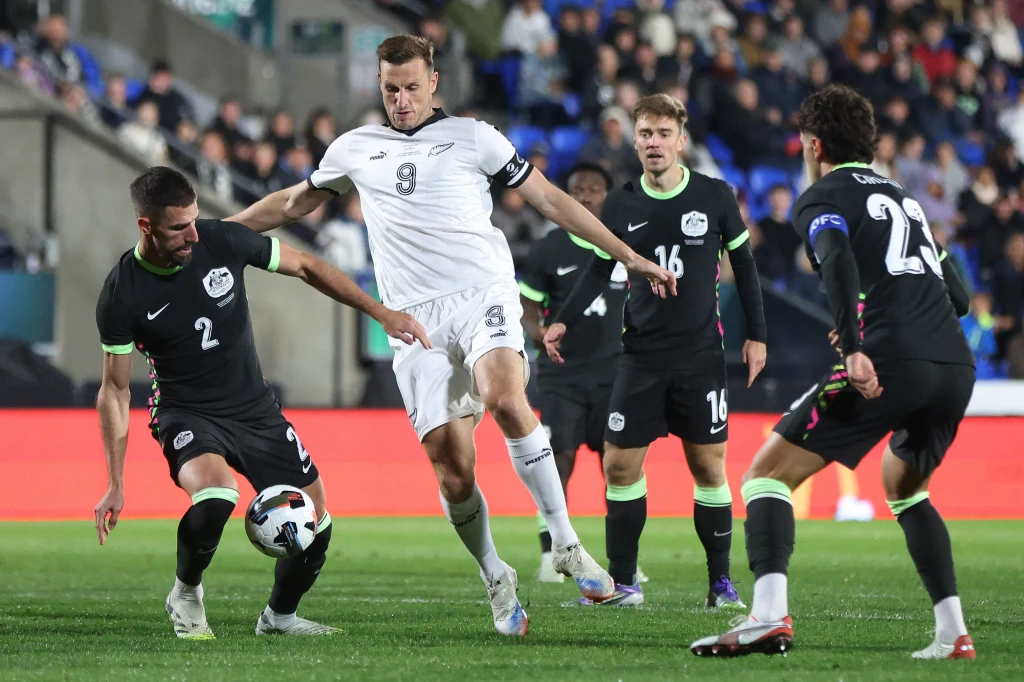 Chris Wood w meczu z reprezentacją Australii Zawodnicy dwóch drużyn piłkarskich walczą o piłkę na murawie stadionu, w tle widoczne są trybuny z kibicami, zawodnicy skupieni są na grze, jeden z nich wykonuje ruch w kierunku piłki, a pozostali obserwują sytuację.