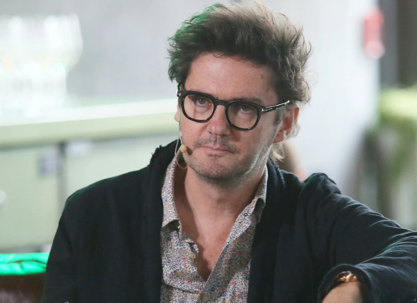 Kuba Wojewódzki Mężczyzna w okularach z mikrofonem na uchu, ubrany w ciemną marynarkę i wzorzystą koszulę, siedzi w jasnym wnętrzu.