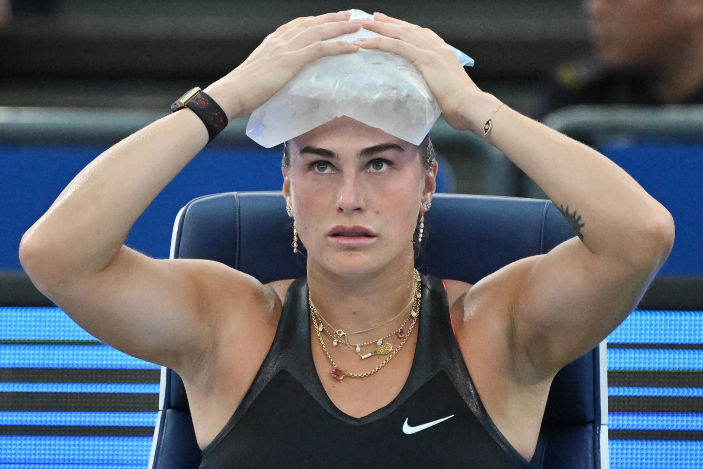 Zawodniczka tenisa w czarnej sportowej koszulce siedzi na krześle, trzymając worek z lodem na głowie. Ma skupiony wyraz twarzy, na szyi nosi złote łańcuszki, a w tle widoczne są rozmyte postacie.