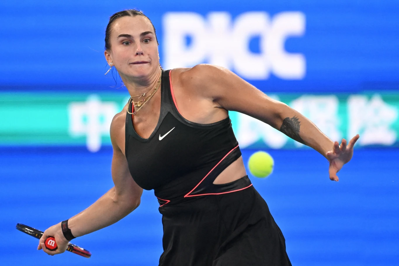 Aryna Sabalenka Tenisistka w czarnej sportowej sukience podczas dynamicznego uderzenia piłki tenisowej na korcie, w tle niebieskie tablice reklamowe.