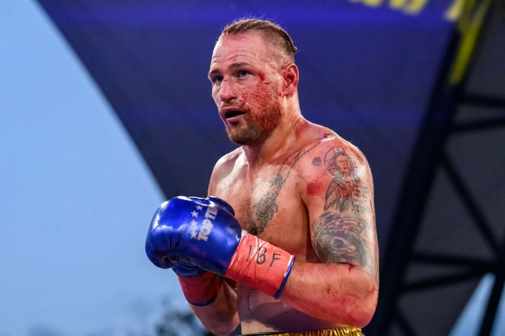 Bokser z licznymi tatuażami, zakrwawioną twarzą i niebieskimi rękawicami stoi na ringu podczas walki. W tle widoczna konstrukcja namiotu i rozmyte światła.