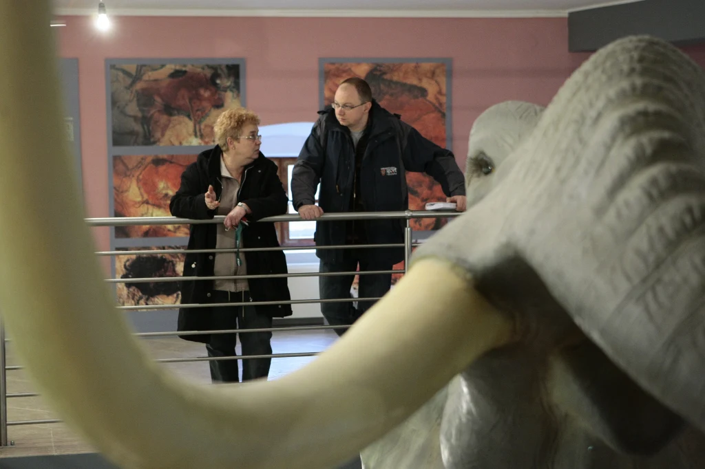 Autor tekstu przy rekonstrukcji Palaeoloxodon antiquus w Muzeum Okręgowym w Koninie Dwie osoby rozmawiające przy balustradzie w muzeum, na pierwszym planie widoczny fragment rekonstrukcji mamuta, w tle ekspozycja z ilustracjami prehistorycznych scen.