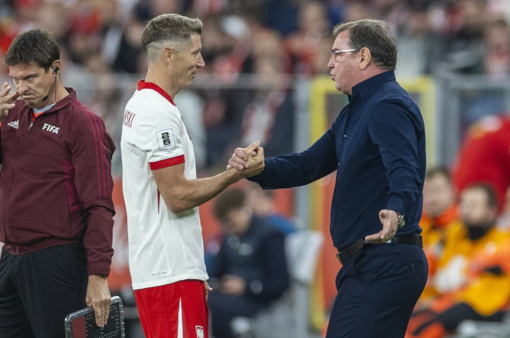 Jan Urban znalazł wspólny język z Robertem Lewandowskim Uścisk dłoni pomiędzy polskim piłkarzem w białej koszulce z czerwonymi akcentami a mężczyzną w niebieskiej koszuli na tle stadionu wypełnionego kibicami.