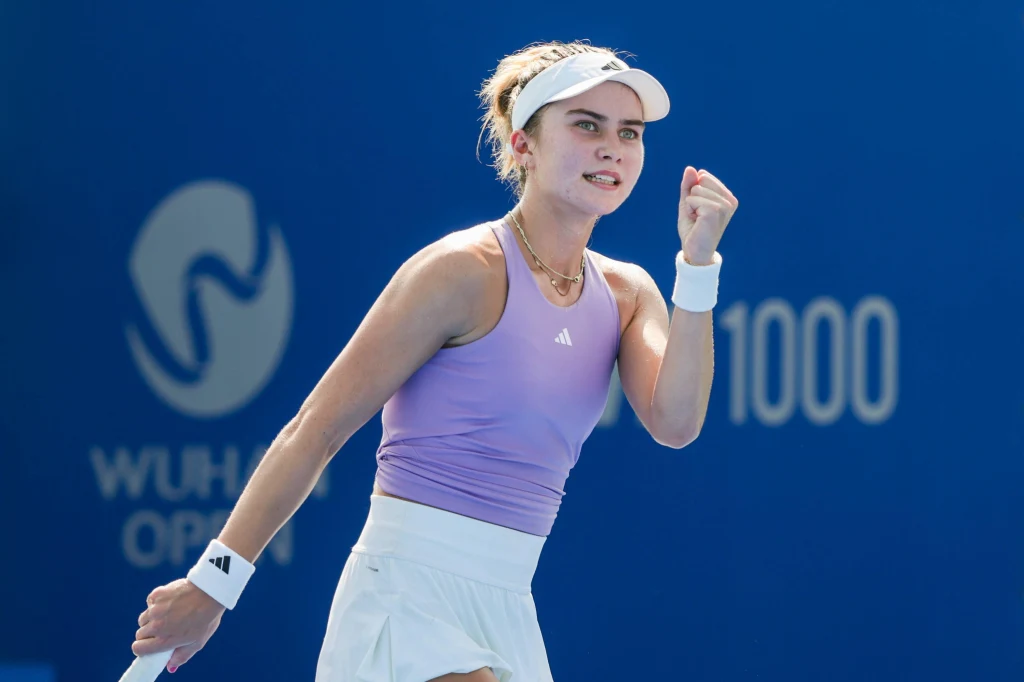 Iva Jovic Zawodniczka tenisowa w sportowym stroju wykonuje gest zwycięstwa z zaciśniętą pięścią na tle niebieskiej tablicy z logo turnieju i napisem WTA 1000.