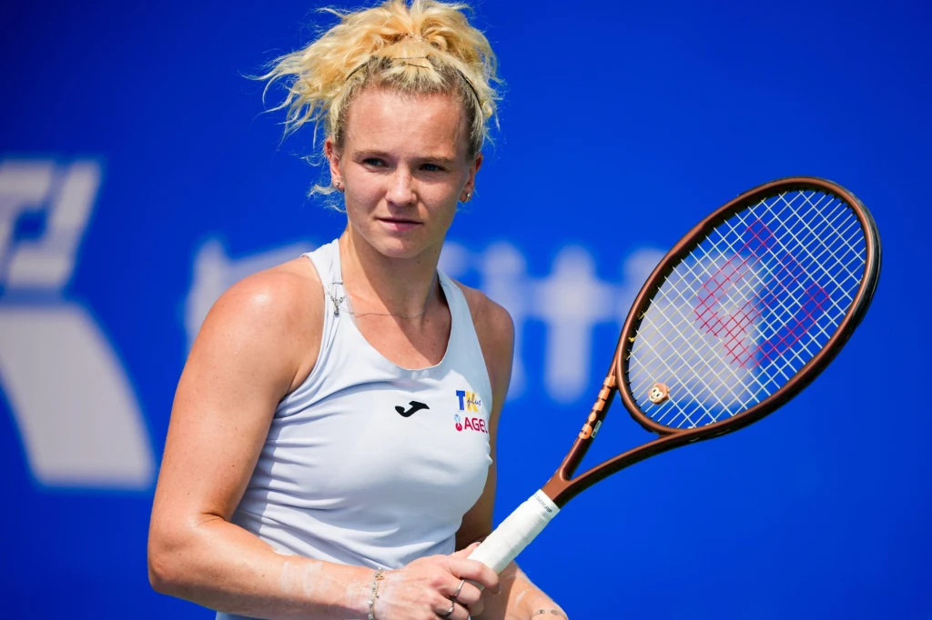 Katerina Siniakova Kobieta w sportowej białej koszulce trzymająca rakietę tenisową na tle niebieskiej ściany, skoncentrowana na grze.