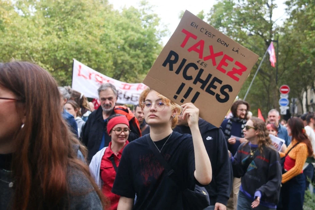 Demonstracja w Paryżu na rzecz opodatkowania najbogatszych Grupa ludzi maszerująca podczas protestu, młoda kobieta na pierwszym planie trzyma kartonowy transparent z napisem 'Taxez les riches', wokół niej widać inne osoby i transparenty, w tle drzewa i zieleń