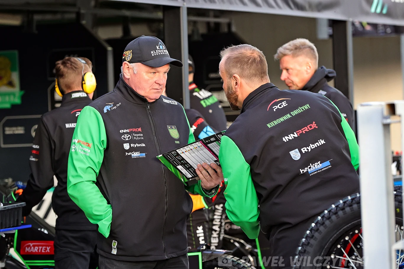 Trener ROW-u Rybnik Piotr Żyto. Mężczyźni w sportowych kurtkach drużynowych rozmawiają w boksie żużlowym, jeden z nich trzyma tablicę z danymi technicznymi; w tle inni członkowie zespołu przygotowują motocykl.