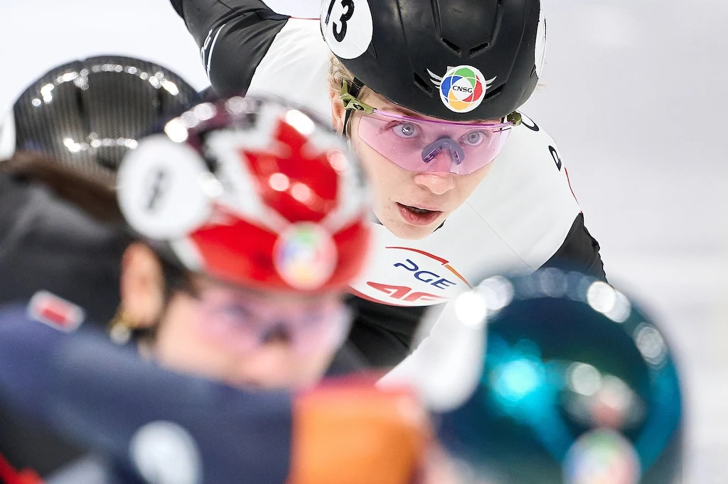 Zawodnik wyścigów na łyżwach podczas sportowej rywalizacji na lodzie, w dynamicznej pozie, z wyraźnym skupieniem na twarzy, widoczne profesjonalne stroje sportowe oraz numer startowy.