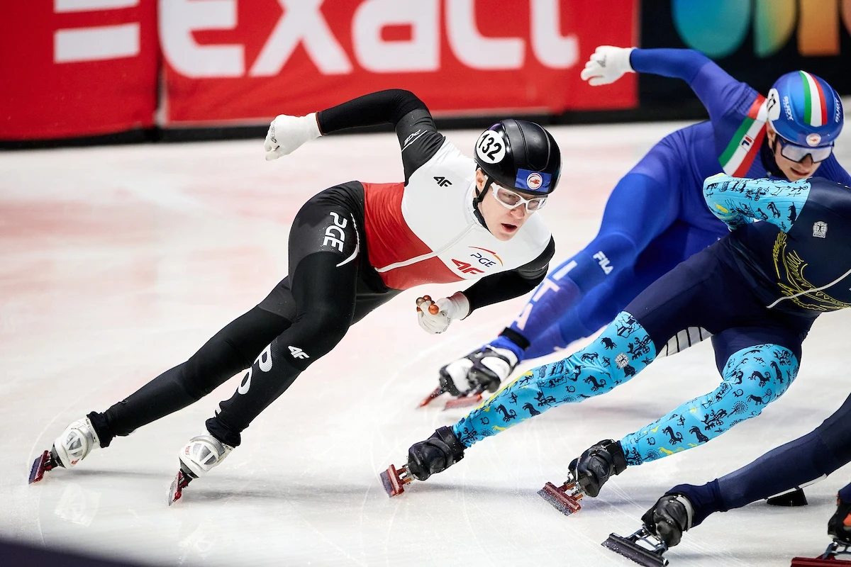 Zawodnicy podczas wyścigu łyżwiarskiego na krótkim torze, pochylają się dynamicznie w zakręcie, ubrani w kombinezony sportowe, ochraniacze i kaski, rywalizacja odbywa się na lodowisku.