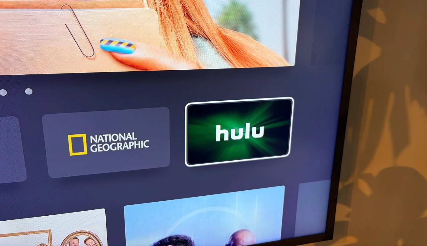 Hulu debiutuje w polskiej wersji Disney+. Jaki ma to wpływ na treści? Ekran telewizora z widocznymi aplikacjami streamingowymi, w tym National Geographic oraz Hulu, z wyraźnymi logotypami na ciemnym tle, w górnej części fragment ręki osoby trzymającej kartonowe pudełko.