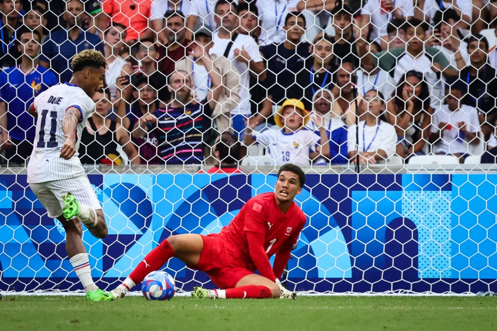 Piłkarz w białym stroju oddaje strzał na bramkę, bramkarz w czerwonym stroju próbuje zatrzymać piłkę, w tle widać tłum kibiców za siatką bramki.