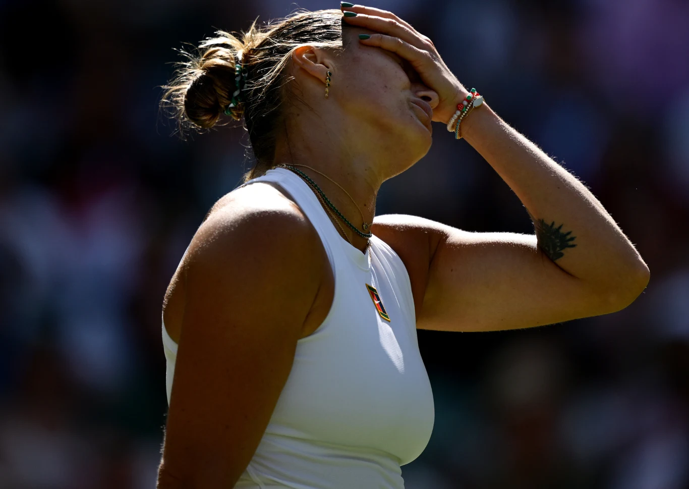 Aryna Sabalenka Zawodniczka w stroju sportowym trzyma dłoń na twarzy, wyrażając emocje związane z rozczarowaniem lub zmęczeniem, silnie oświetlona przez promień światła, w tle zamazany stadion.