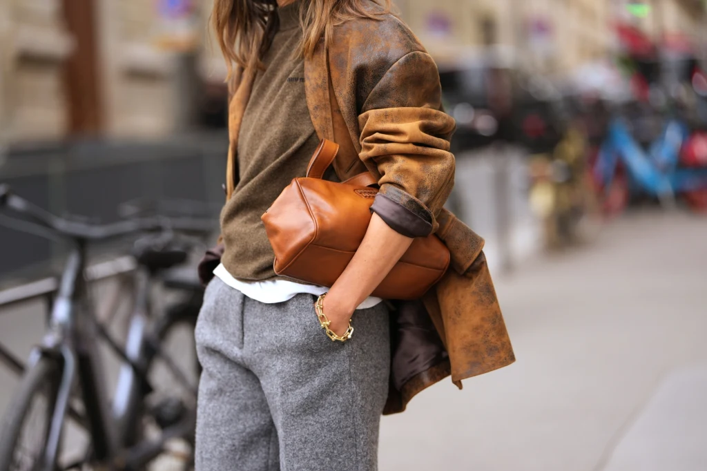 Praktyczna moda na jesień Stylowa kobieta w brązowej skórzanej kurtce, oliwkowym swetrze i szarych, wełnianych spodniach trzyma w ręce elegancką brązową torebkę na tle miejskiej ulicy.