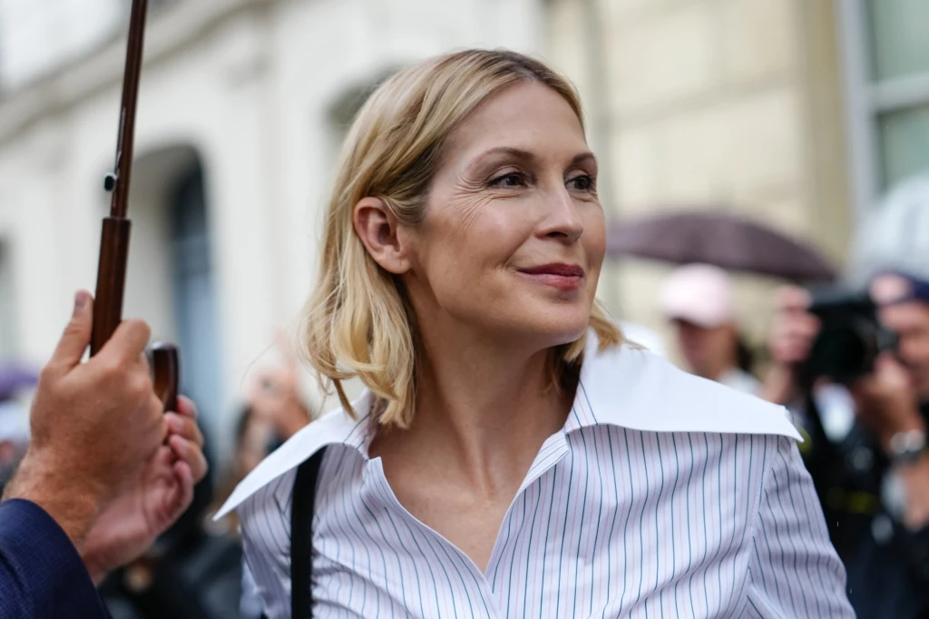 Modowe triki na jesień Uśmiechnięta kobieta w jasnej koszuli z szerokim kołnierzem stoi na tle tłumu i budynków, w otoczeniu kilku osób trzymających parasole.