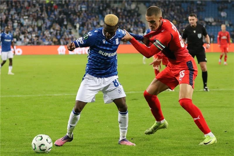 Dwóch piłkarzy rywalizujących o piłkę podczas meczu piłkarskiego na stadionie, jeden ubrany w niebieski strój, drugi w czerwony, w tle widoczni kibice na trybunach.