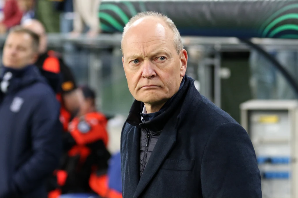 Niels Frederiksen, trener Lecha Poznań Starszy mężczyzna w eleganckim, ciemnym płaszczu stoi na tle stadionu, z poważnym wyrazem twarzy. W tle widać rozmyte postacie, prawdopodobnie członków ekipy technicznej lub ochrony, oraz fragment ławki rezerwowych.