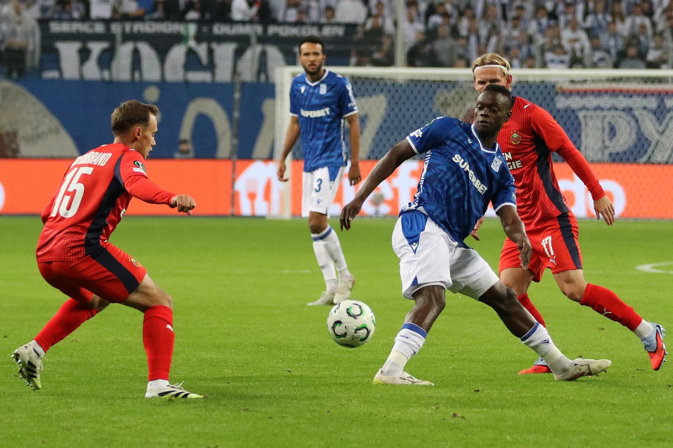 piłkarze w niebieskich i czerwonych strojach rywalizują o piłkę na zielonej murawie stadionu przed licznie zgromadzoną publicznością