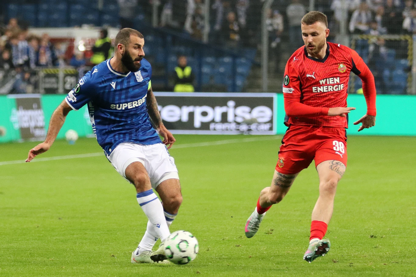 Dwóch piłkarzy w trakcie intensywnej gry na stadionie, jeden ubrany w niebieski strój drużyny Lech Poznań, drugi w czerwonym stroju drużyny przeciwnika, na zielonej murawie, w tle trybuny z kibicami.