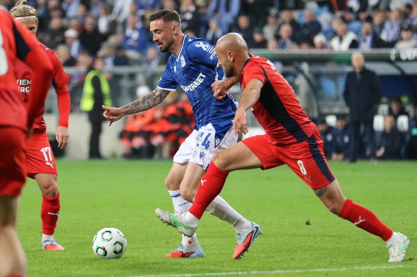 Dwóch piłkarzy w akcji podczas meczu piłkarskiego, jeden ubrany w niebieski strój prowadzi piłkę, drugi w czerwonym próbuje ją odebrać, tłum kibiców widoczny w tle.