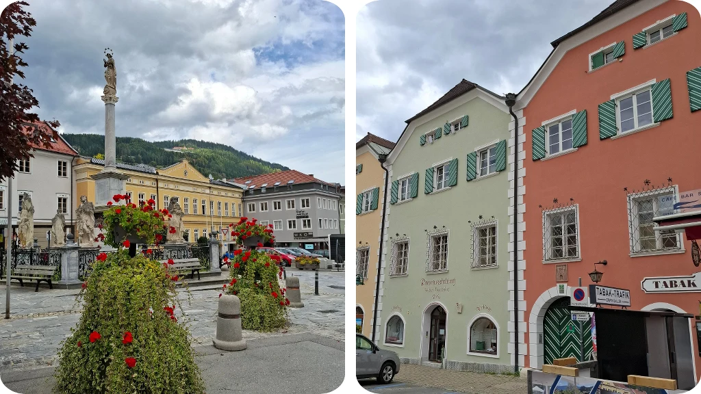 Rynek miejski z kolorowymi donicami z kwitnącymi kwiatami oraz kolumną pośrodku, w tle pastelowe kamienice oraz górski krajobraz z zielonymi wzgórzami i częściowo zachmurzonym niebem.