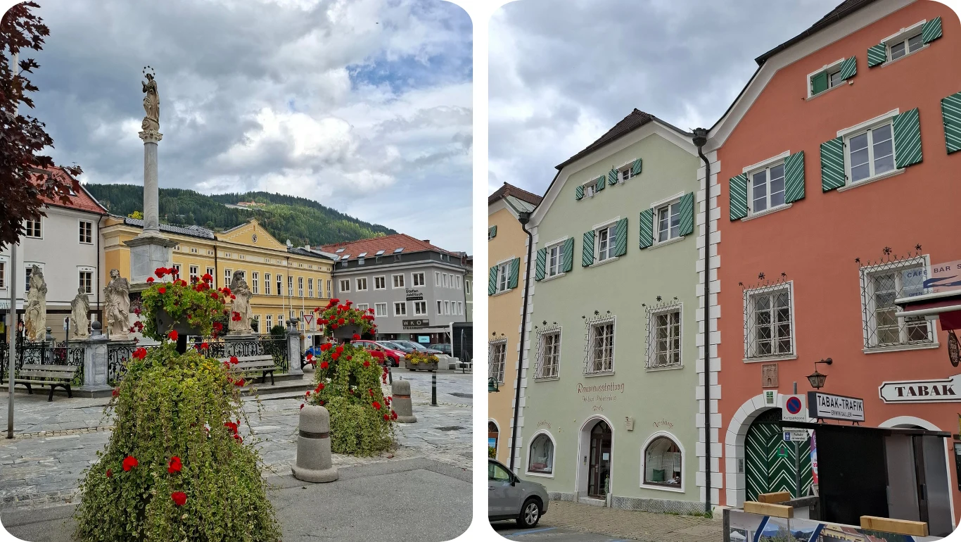 Rynek w Murau Rynek miejski z kolorowymi donicami z kwitnącymi kwiatami oraz kolumną pośrodku, w tle pastelowe kamienice oraz górski krajobraz z zielonymi wzgórzami i częściowo zachmurzonym niebem.