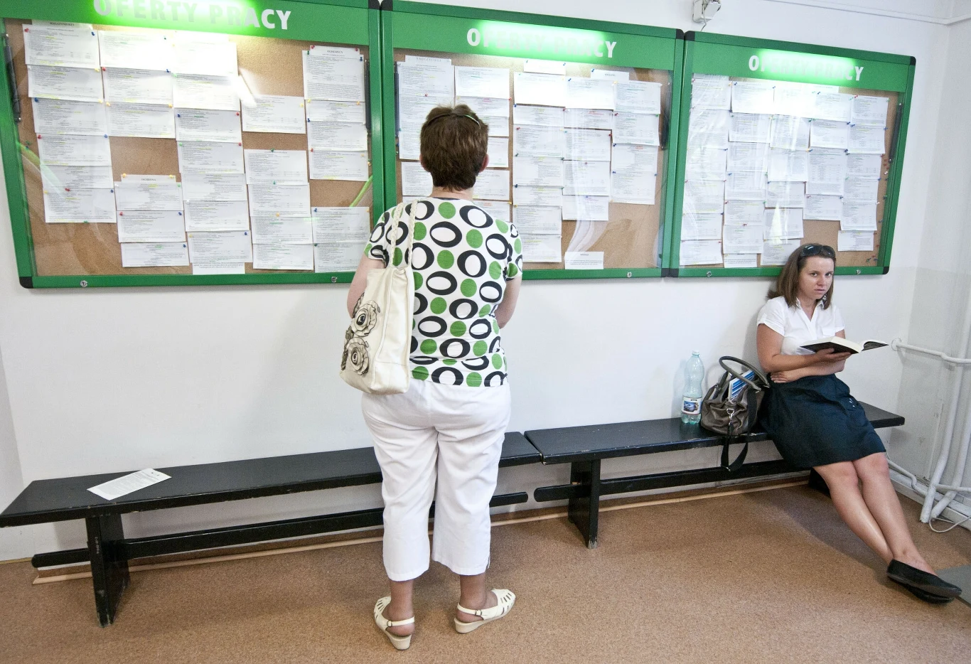 Od czerwca w Polsce stopa bezrobocia rośnie, choć wciąż pozostaje niska na tle krajów UE. Jaka jest kondycja krajowego rynku pracy? Kobieta w białych spodniach i bluzce w kolorowe kropki stoi przed dużą tablicą ogłoszeń zapełnioną kartkami, obok na ławce siedzi kobieta czytająca książkę. Tablica z nagłówkiem 'oferty pracy' dominuje w pomieszczeniu.