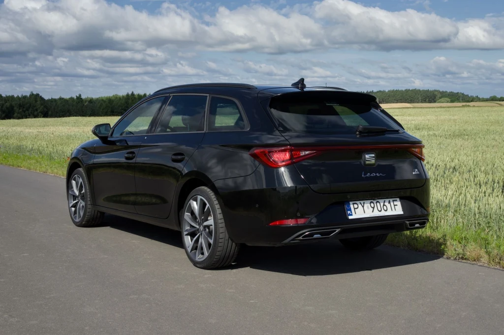 Seat Leon po liftingu, w przeciwieństwie do Cupry Leon, nie uległ zmianom stylistycznym. W ten sposób Hiszpanie wprowadzili rozróżnienie między modelem oferowanym pod tymi dwiema markami. Czarny samochód Seat Leon ST stoi zaparkowany na asfaltowej drodze prowadzącej przez zielone pole, w tle widoczne są drzewa i niebo z chmurami.