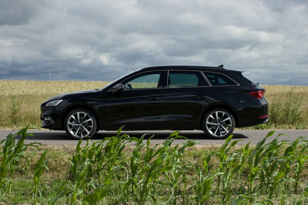 Seat Leon e-Hybrid, podobnie jak przed modernizacją generuje 204 KM, ale został wyposażony w większy silnik benzynowy (1.5 zamiast 1.4) i większy akumulator (19,7 kWh netto). Czarny samochód kombi zaparkowany na poboczu drogi, w tle pole zboża i pochmurne niebo, na pierwszym planie fragment zielonej roślinności.