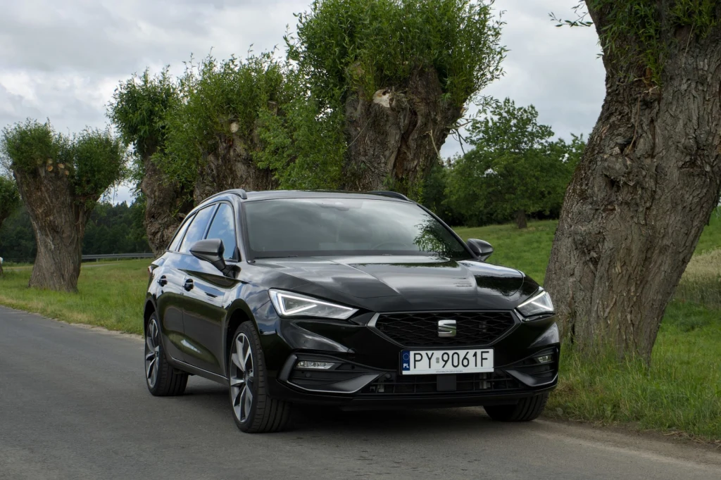 Seat Leon e-Hybrid z nową hybrydą plug-in rozpędza się do 100 km/h o 0,2 s wolniej niż przed liftingiem. Czarny samochód osobowy marki Seat stoi zaparkowany na poboczu asfaltowej drogi, otoczony rzędem przyciętych wierzb i zielonym krajobrazem wiejskim, pod pochmurnym niebem.