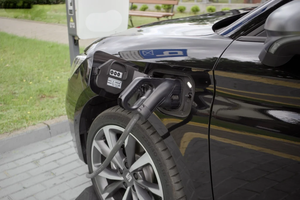 Seat deklaruje, że Leon Sportstourer e-Hybrid może przejechać na prądzie nawet 131 km w cyklu mieszanym. Czarny samochód elektryczny podłączony do publicznej stacji ładowania, widoczny przewód ładujący oraz fragment felgi, w tle fragment chodnika i zieleń.