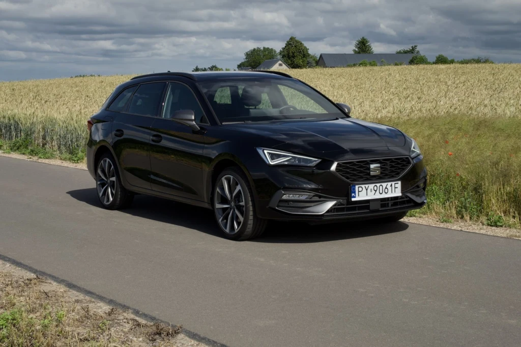 Za Seata Leona Sportstourer e-Hybrid z roku modelowego 2026 trzeba dziś zapłacić co najmniej 173 600 zł. Czarny samochód osobowy marki SEAT parkuje na wąskiej, asfaltowej drodze otoczonej polami zboża pod zachmurzonym niebem.