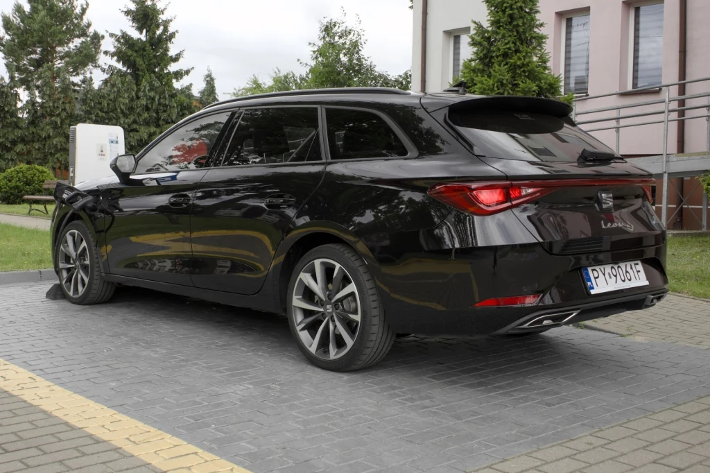 Seat Leon e-Hybrid obsługuje ładowanie prądem stałym o mocy 40 kW. Czarny samochód typu kombi marki SEAT Leon zaparkowany na kostce brukowej przy budynku z jasnymi ścianami, w tle widoczne drzewa oraz krzewy.