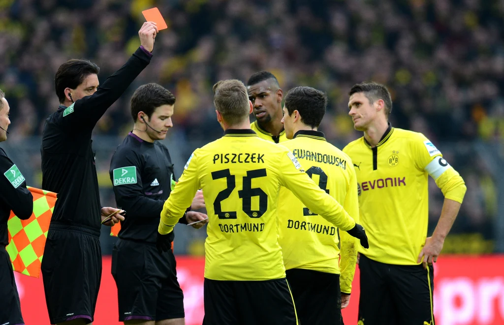 Sędzia wręcza czerwoną kartkę piłkarzowi podczas meczu, otoczony przez kilku zawodników w żółto-czarnych strojach drużyny Borussia Dortmund, tłum kibiców widoczny w tle.