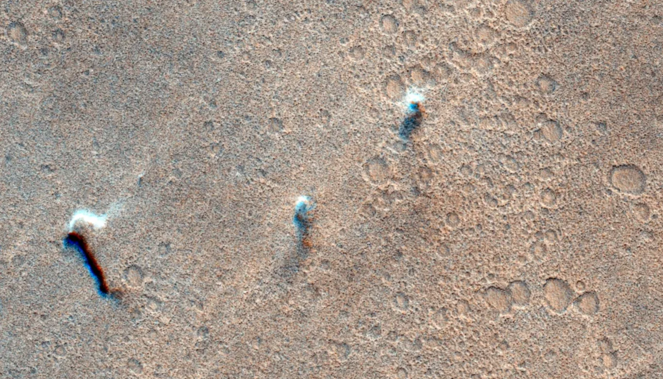 Trzy wiry pyłowe przemieszczające się po powierzchni Marsa 8 listopada 2021 roku Powierzchnia pokryta licznymi, niewielkimi kraterami oraz trzema wzniesieniami rzucającymi długie cienie na piaskowe, marsjańskie podłoże.