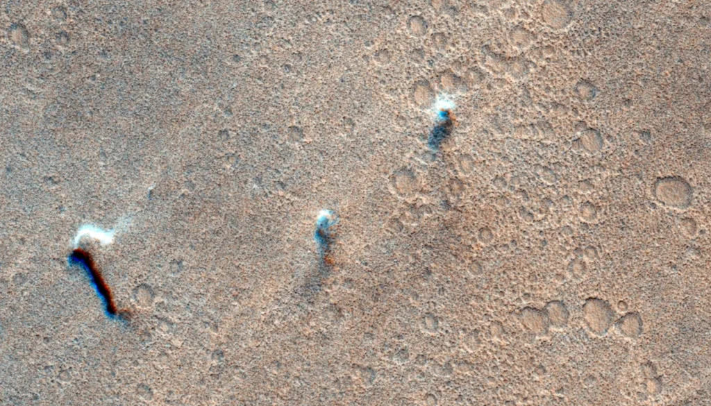 Trzy wiry pyłowe przemieszczające się po powierzchni Marsa 8 listopada 2021 roku Powierzchnia pokryta licznymi, niewielkimi kraterami oraz trzema wzniesieniami rzucającymi długie cienie na piaskowe, marsjańskie podłoże.