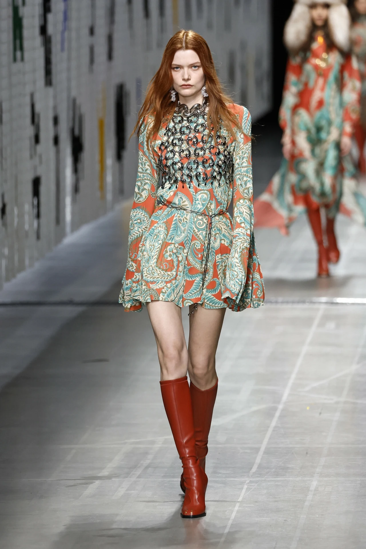 Modelka w kolorowej sukience o wzorze paisley z długimi rękawami i czerwonych kozaczkach idzie po wybiegu, w tle druga osoba w podobnej stylizacji, scena jasna i nowoczesna.