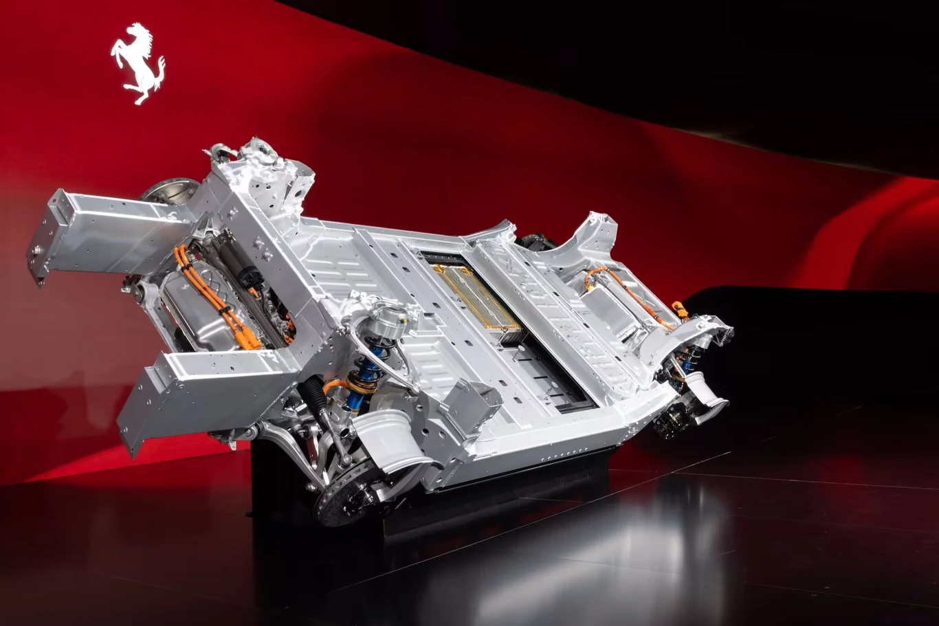 Podwozie elektrycznego samochodu Ferrari wyeksponowane na tle czerwonej ściany z charakterystycznym logo marki, widoczne elementy konstrukcyjne, akumulatory i przewody elektryczne.