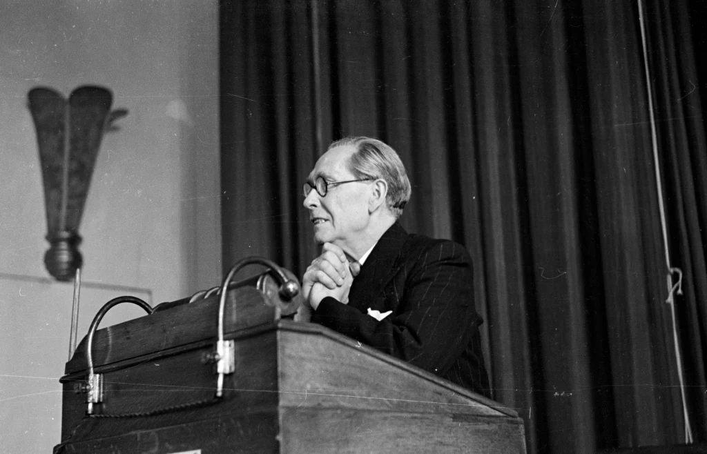 Starszy mężczyzna w garniturze mówi przy mównicy na tle zasłoniętej kotary, skupiony na swoim przemówieniu.