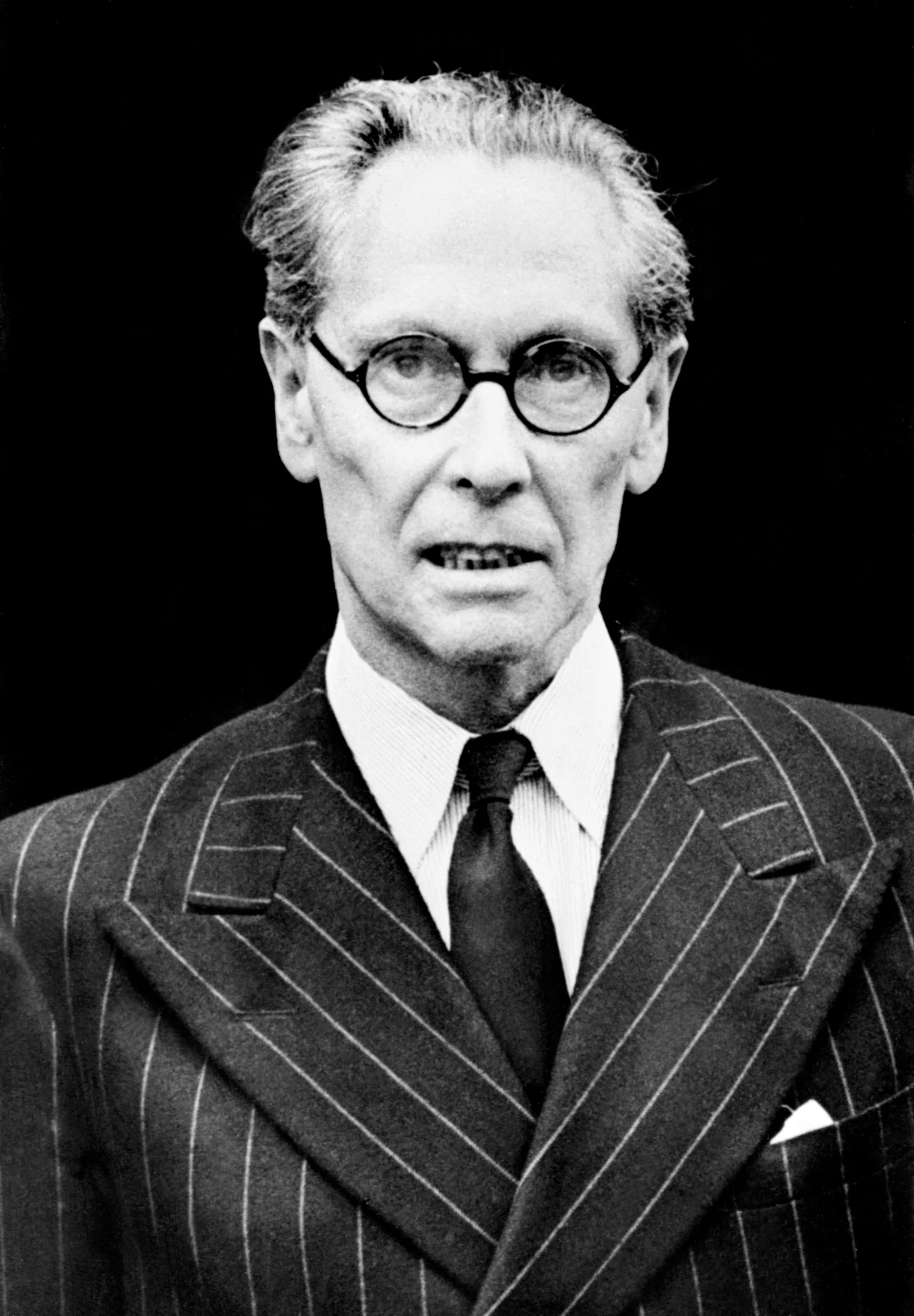 Starszy mężczyzna w okularach, w eleganckim, prążkowanym garniturze z białą koszulą i krawatem, patrzy poważnie w obiektyw. Tło jest całkowicie czarne, co podkreśla postać.