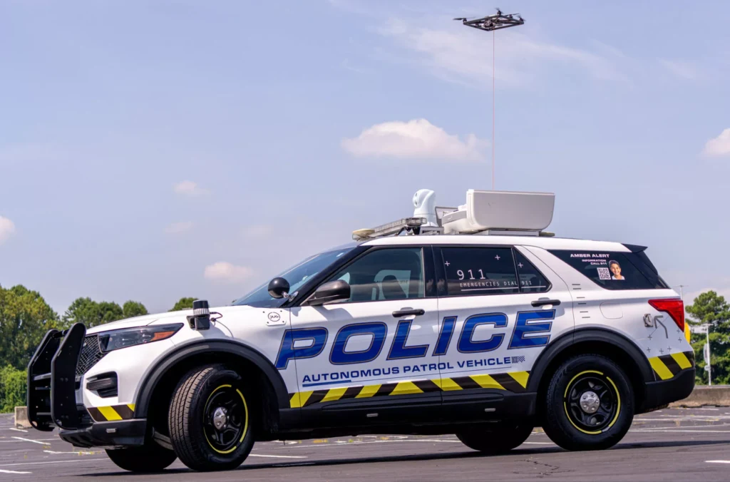 PUG Patrol Partner – sztuczna inteligencja patroluje ulice. Autonomiczny radiowóz policyjny zaparkowany na asfalcie, z widoczną sygnalizacją i żółto-czarnymi pasami ostrzegawczymi na bokach, nad pojazdem zawieszony dron patrolujący teren