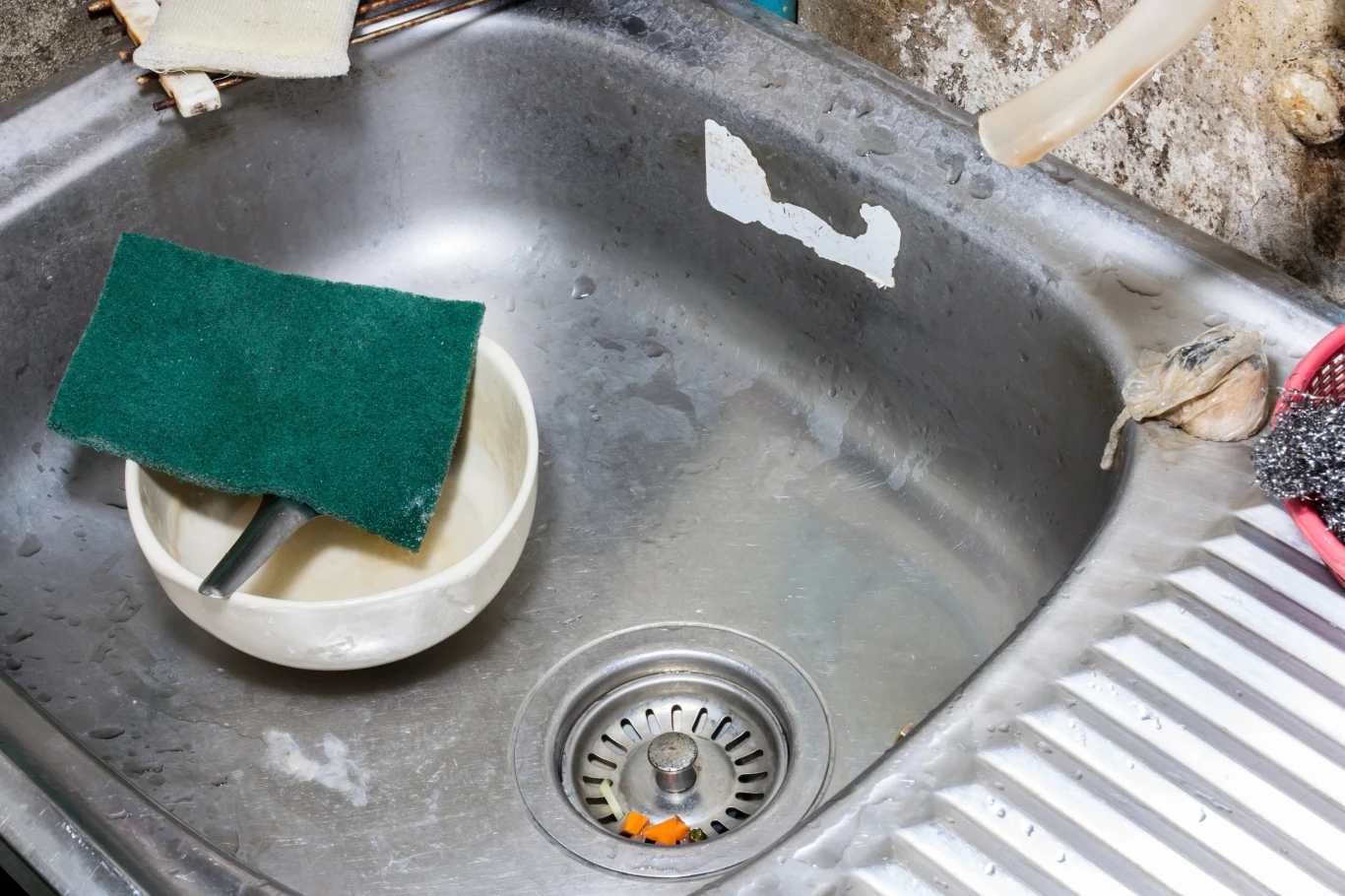 Poznaj najczęstsze błędy, przez które zatyka się odpływ w kuchni Zardzewiały zlew kuchenny z odpływem, w środku umieszczona plastikowa miska z gąbką do mycia naczyń, ślady zabrudzeń, zniszczenie na krawędziach i ścianie wokół zlewu, fragment metalowego ociekacza po prawej stronie.