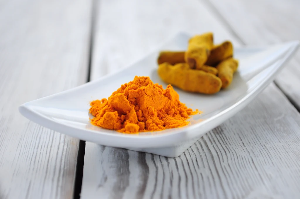Kopczyk intensywnie żółtego proszku kurkumy na białym, prostokątnym talerzyku, z tyłu kilka kawałków korzenia kurkumy, całość ustawiona na jasnym, drewnianym stole.